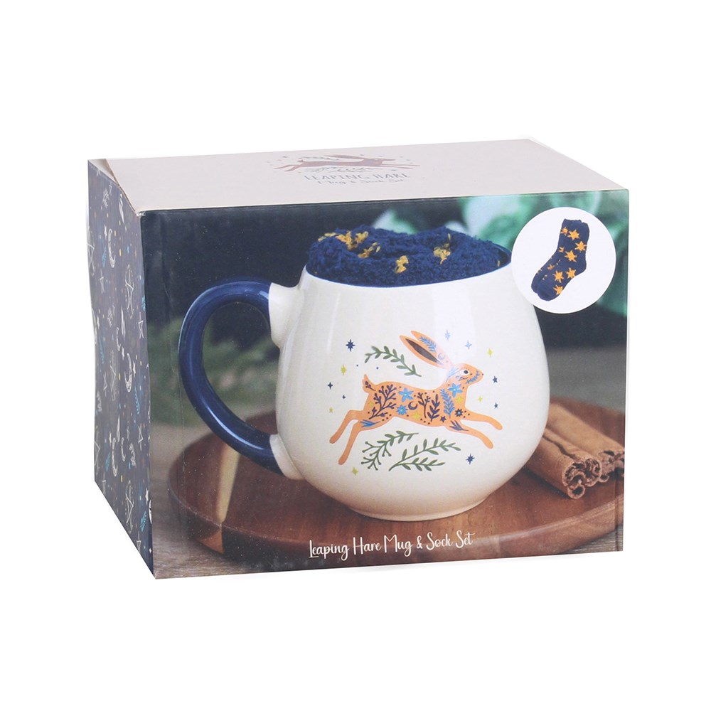 A cream mug with dark blue handle and a picture of a leaping hare with greenery and stars around it. Inside the mug is a folded pair of fluffy dark blue socks with yellow stars. all within the box