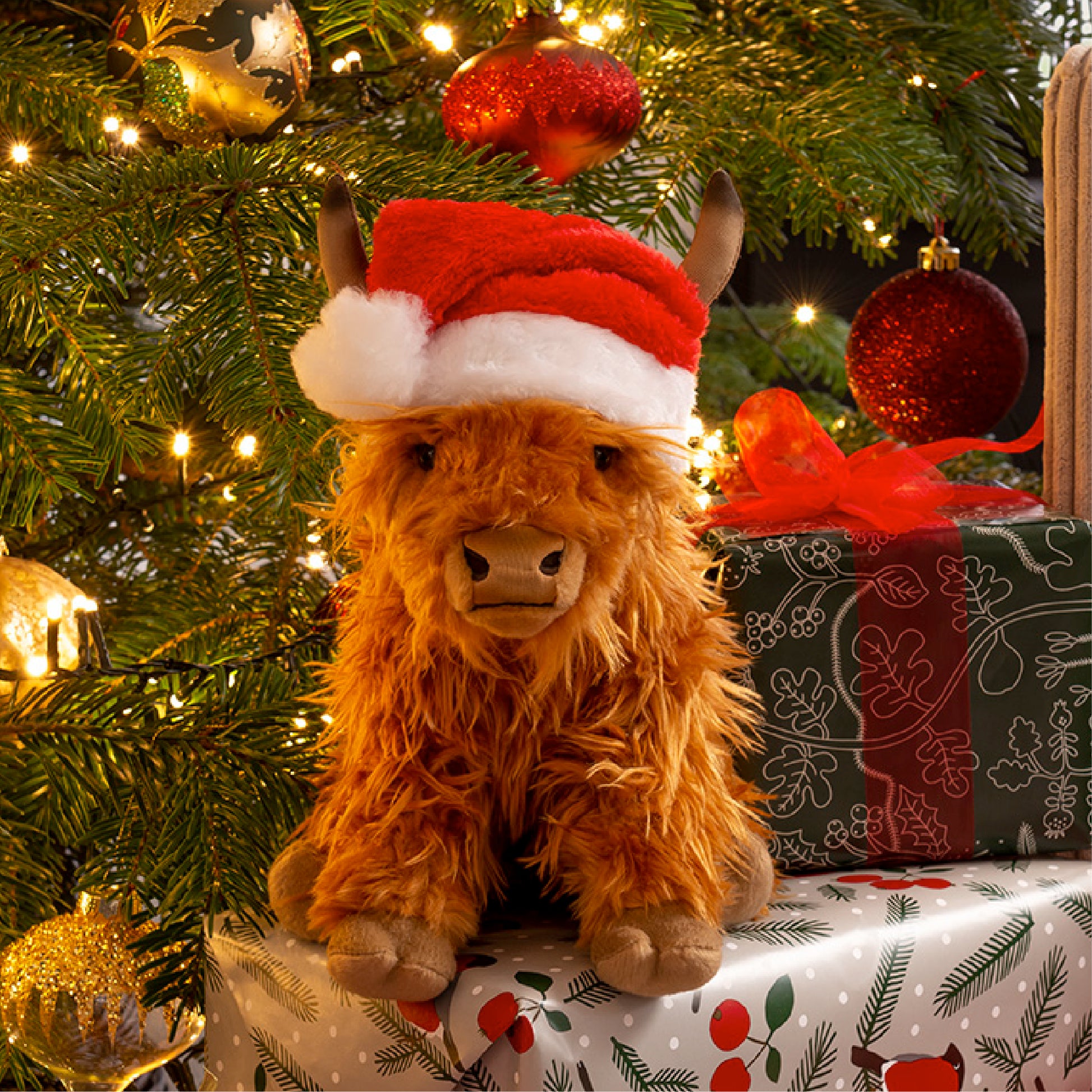 Plush toy cow wearing a Santa hat in front of a decorated Christmas tree with lights and ornaments.