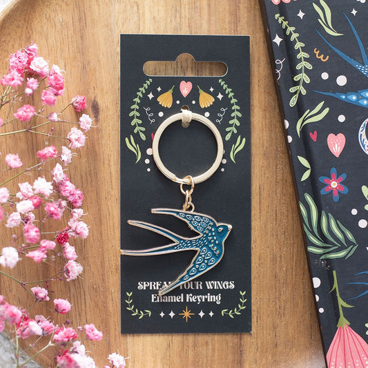 Keyring with blue bird design on a decorative card with floral elements, placed on a wooden surface with pink flowers.