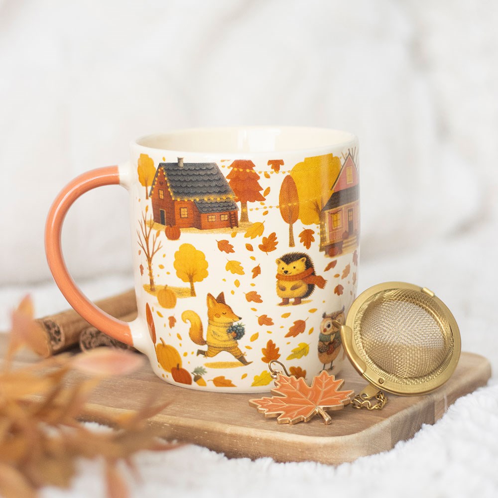 Mug with autumn-themed design featuring houses, trees, and animals on a wooden coaster with a tea ball.