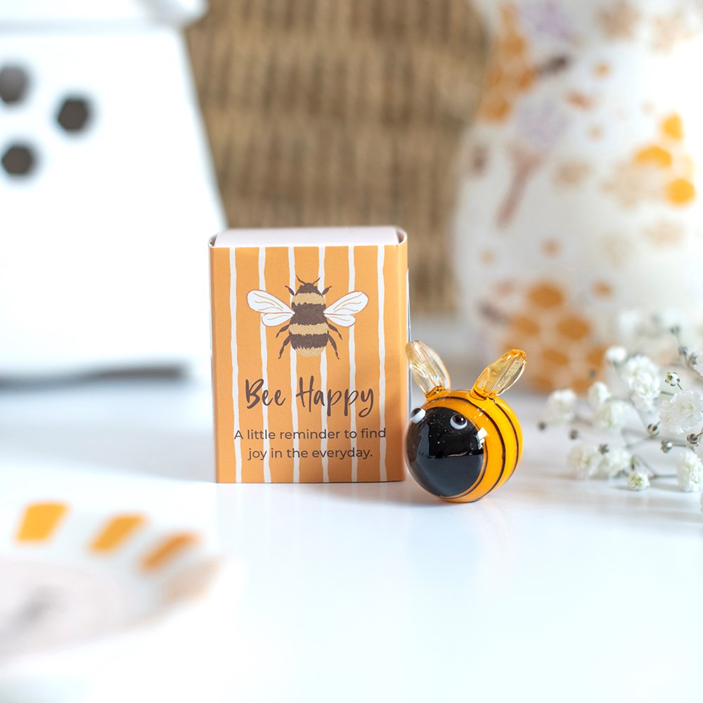 A small bee-shaped glass ornament with a black and yellow striped body and clear wings, placed beside its orange-and-white striped box printed with a bee illustration and the words 'Bee Happy'. The scene is softly lit on a white surface with blurred decorative objects in the background.