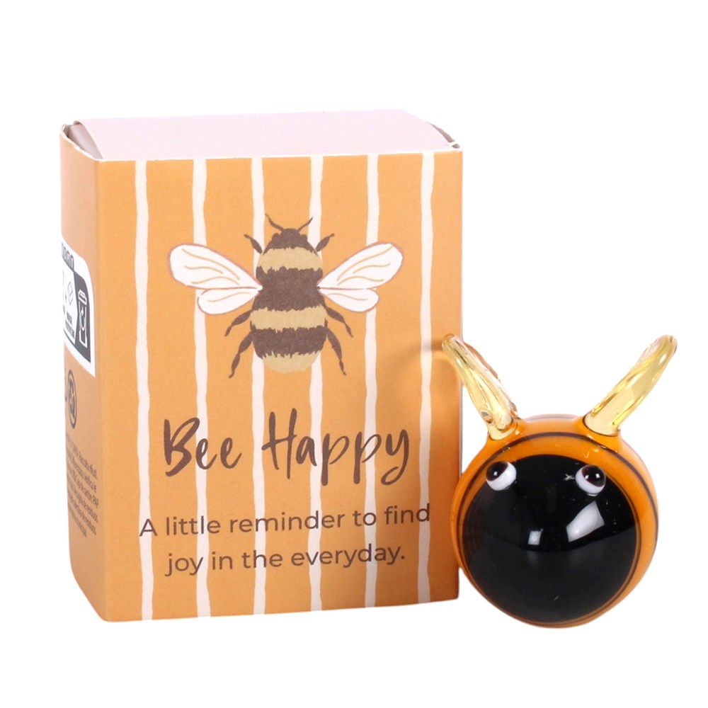 A bee-shaped decorative object with a glossy black face and golden antennae, shown next to its orange box with white stripes and a bee illustration. The box reads 'Bee Happy' and 'A little reminder to find joy in the everyday.