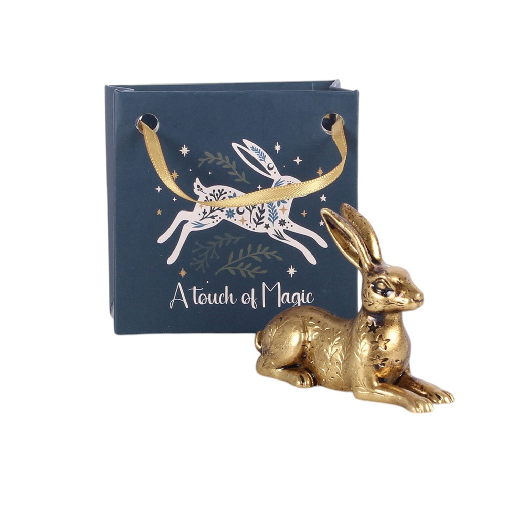 A metallic gold hare ornament positioned in front of a navy blue gift bag featuring a white hare illustration, green leaves, and the text 'A touch of Magic' on a white background.