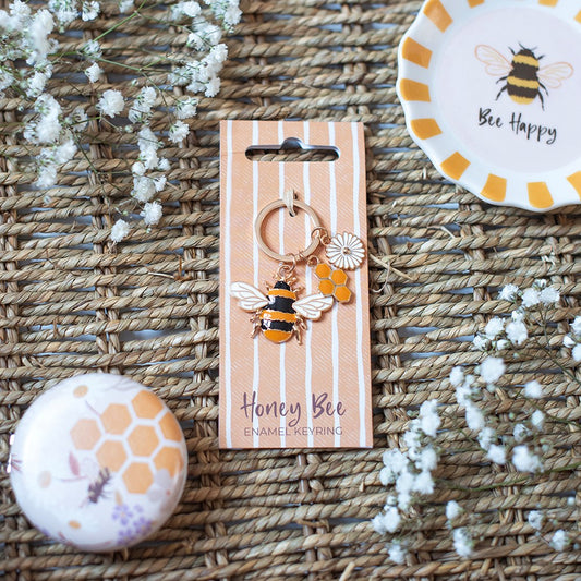 A Honey Bee enamel keyring with a bumblebee, honeycomb, and daisy charm, displayed on its striped cardboard packaging card. The card is styled on a woven straw mat with decorative white flowers and a matching honeycomb pin.