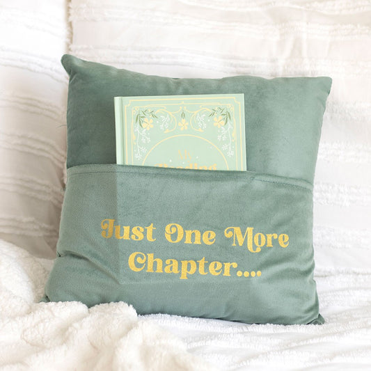 Green pillow with 'Just One More Chapter...' text and a book on a white surface