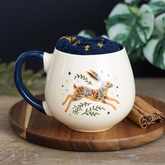 A cream mug with dark blue handle and a picture of a leaping hare with greenery and stars around it. Inside the mug is a folded pair of fluffy dark blue socks with yellow stars.
