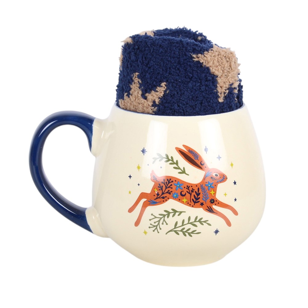 A cream mug with dark blue handle and a picture of a leaping hare with greenery and stars around it. Inside the mug is a folded pair of fluffy dark blue socks with yellow stars.