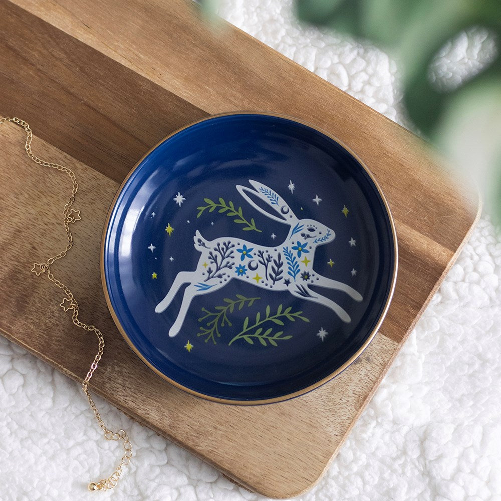 A round ceramic plate with a deep navy blue glaze, featuring a white rabbit decorated with blue and yellow floral patterns, green leaves, and small yellow stars. The plate is styled on a wooden cutting board next to a gold chain necklace on a white textured blanket.