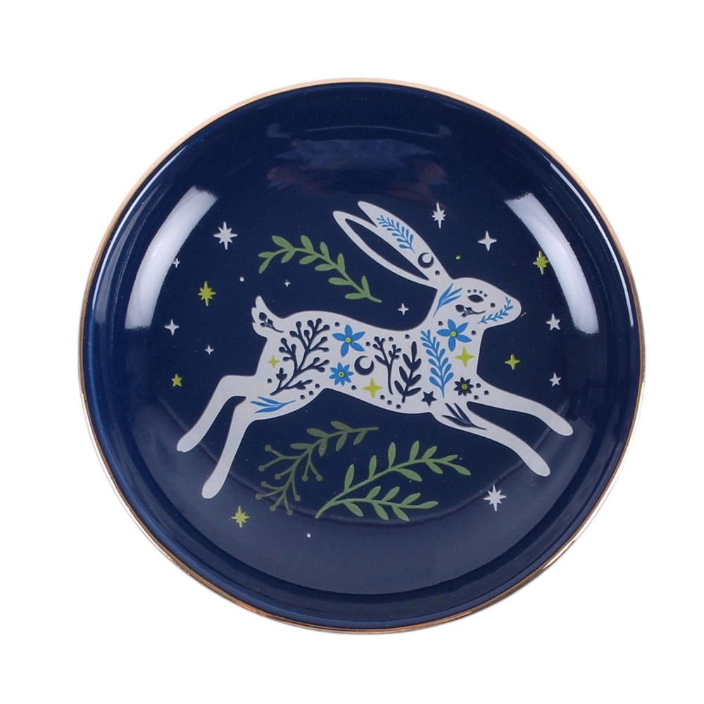 A round navy blue trinket dish featuring a white leaping hare decorated with blue and green floral patterns, surrounded by small white stars and green leafy branches, with a thin gold rim.