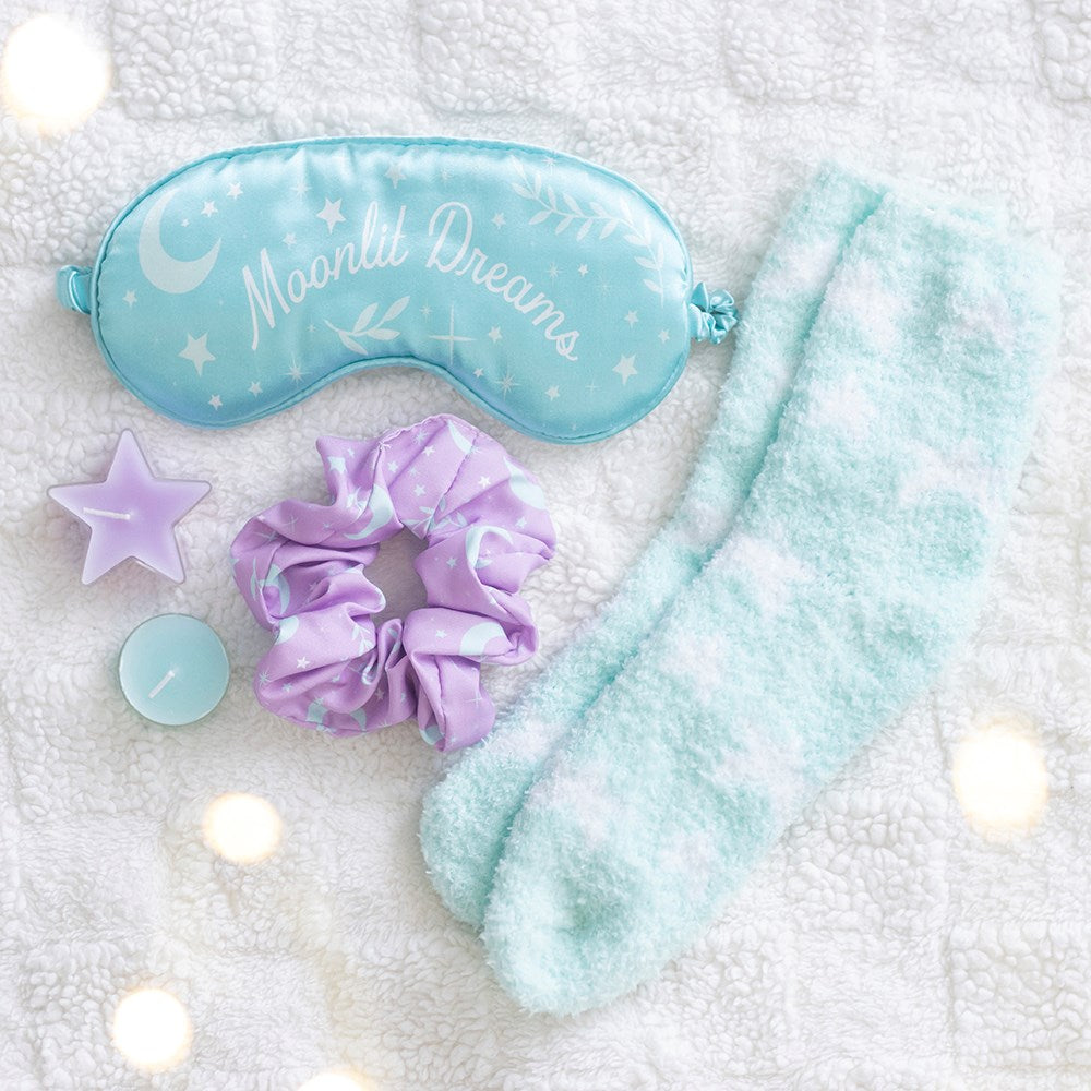 A cozy sleep-themed gift set arranged on a soft white textured blanket with warm bokeh lights. The set includes a light blue satin sleep mask printed with 'Moonlit Dreams' and celestial motifs, fluffy pale blue socks, a lavender satin scrunchie, a lavender star-shaped candle, and a small blue candle.