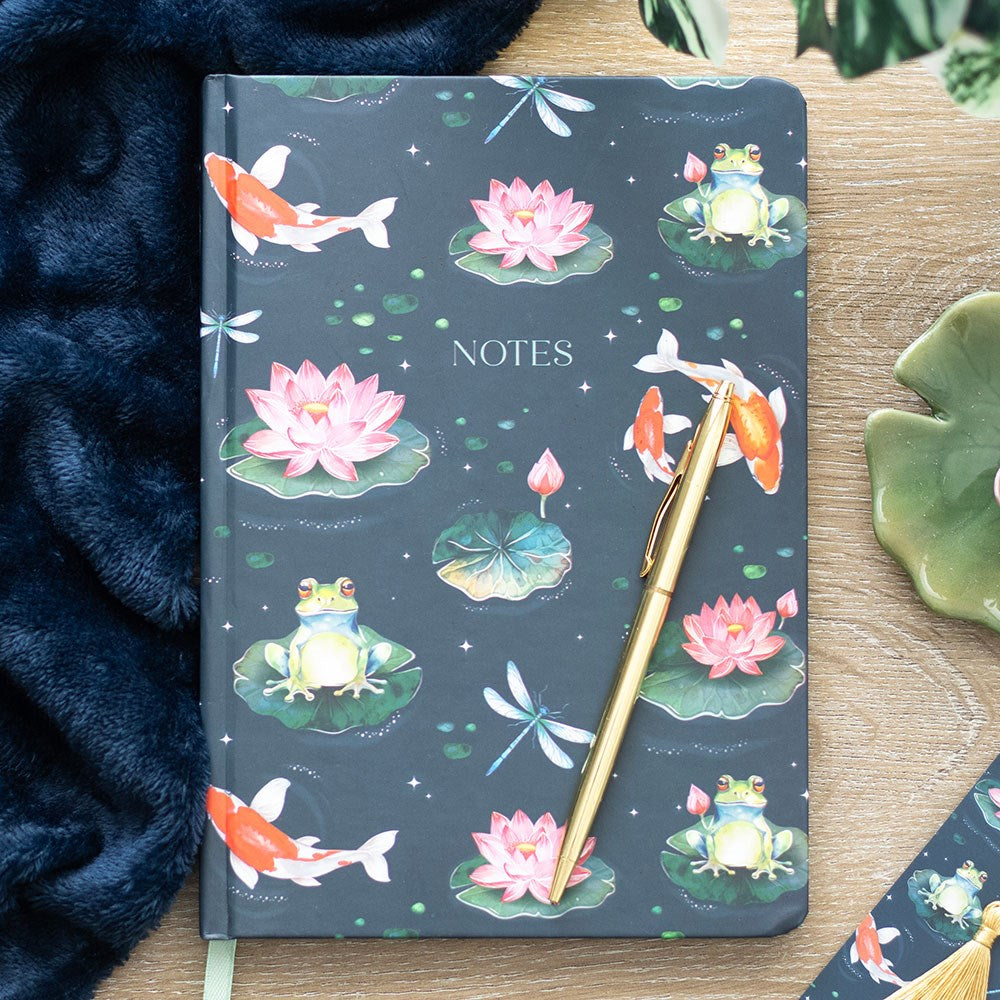 Notebook with pond-themed cover featuring frogs, lotus flowers, and koi fish, accompanied by a gold pen on a wooden surface.