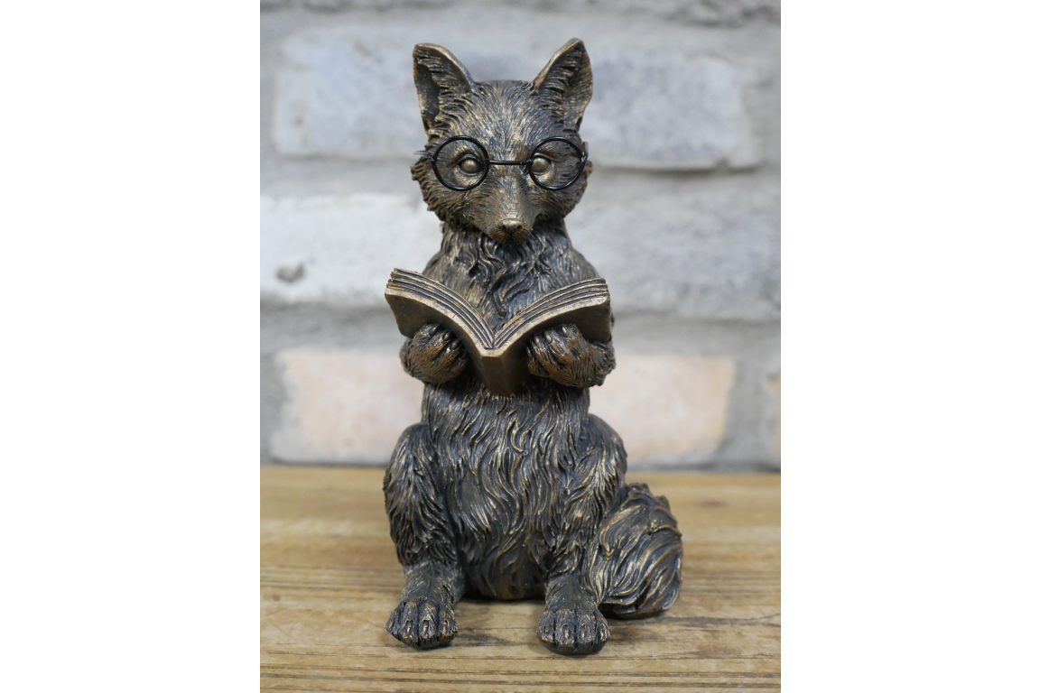 Statue of a fox wearing glasses and reading a book on a wooden surface with a stone wall background.