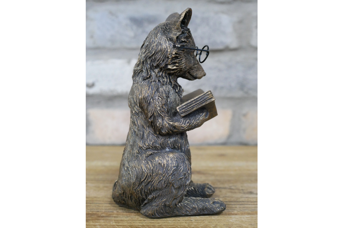 Bronze statue of a fox holding a book on a wooden surface with a stone wall background