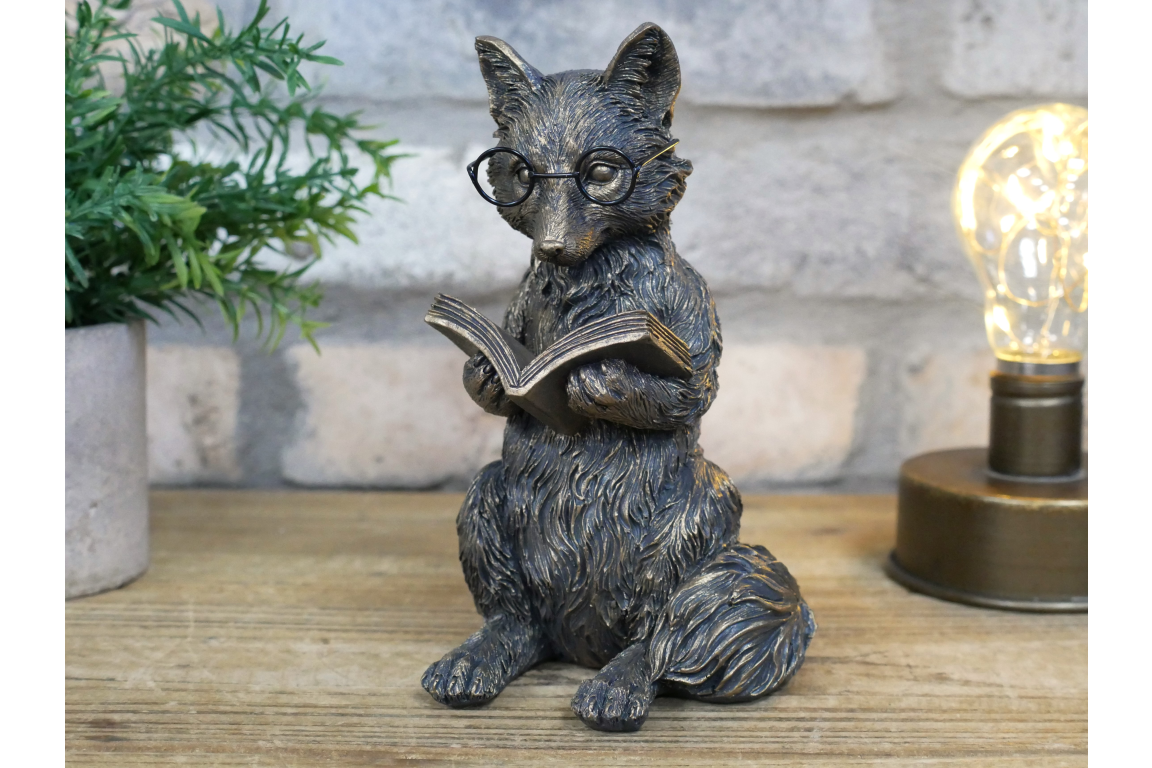 Statue of a fox reading a book with glasses on a wooden surface.