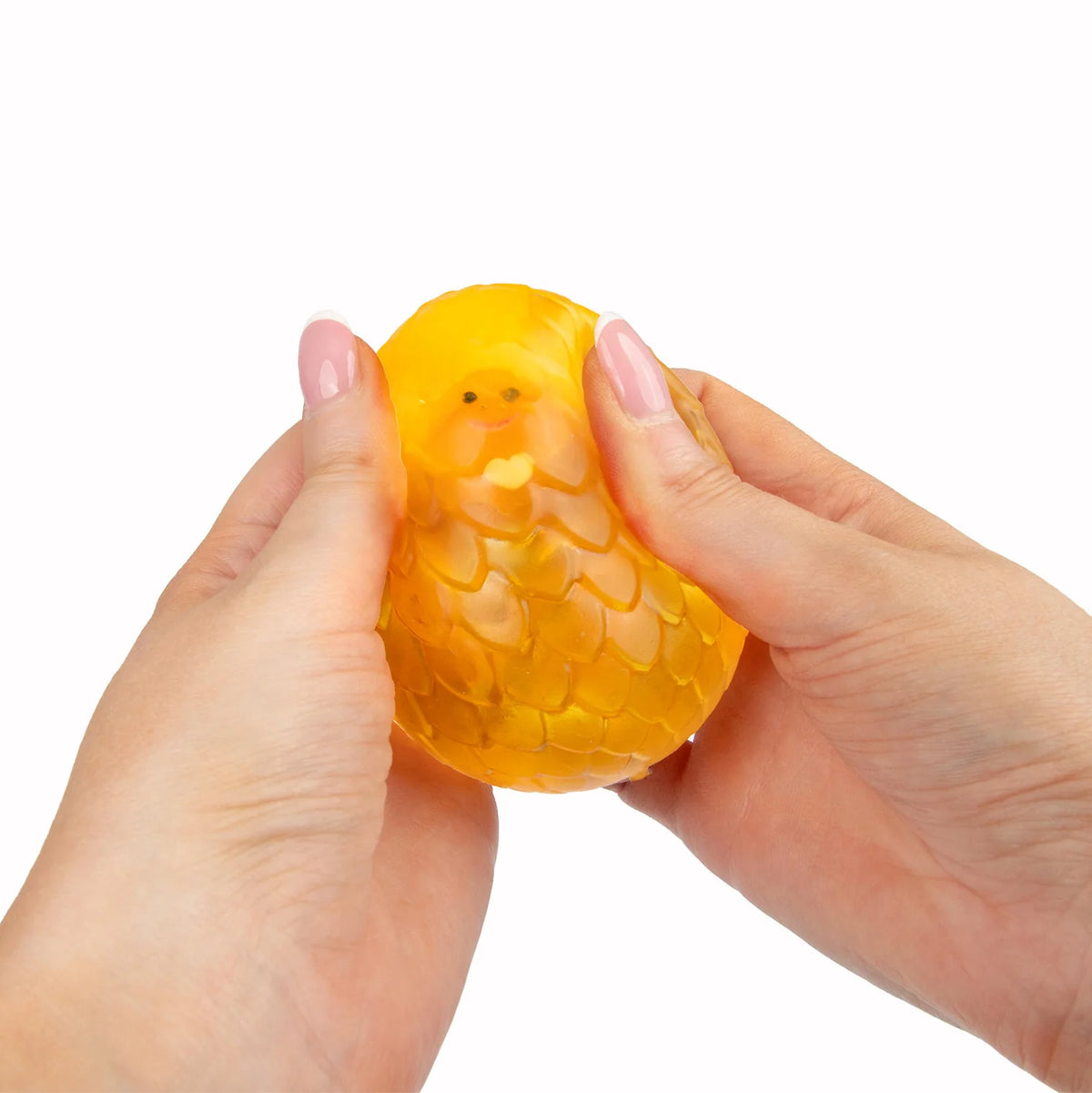 A person's hands squeezing a small, yellow, Scrunchems Sugar Dragon Egg squishy toy with a textured, scale-like surface against a solid white background.