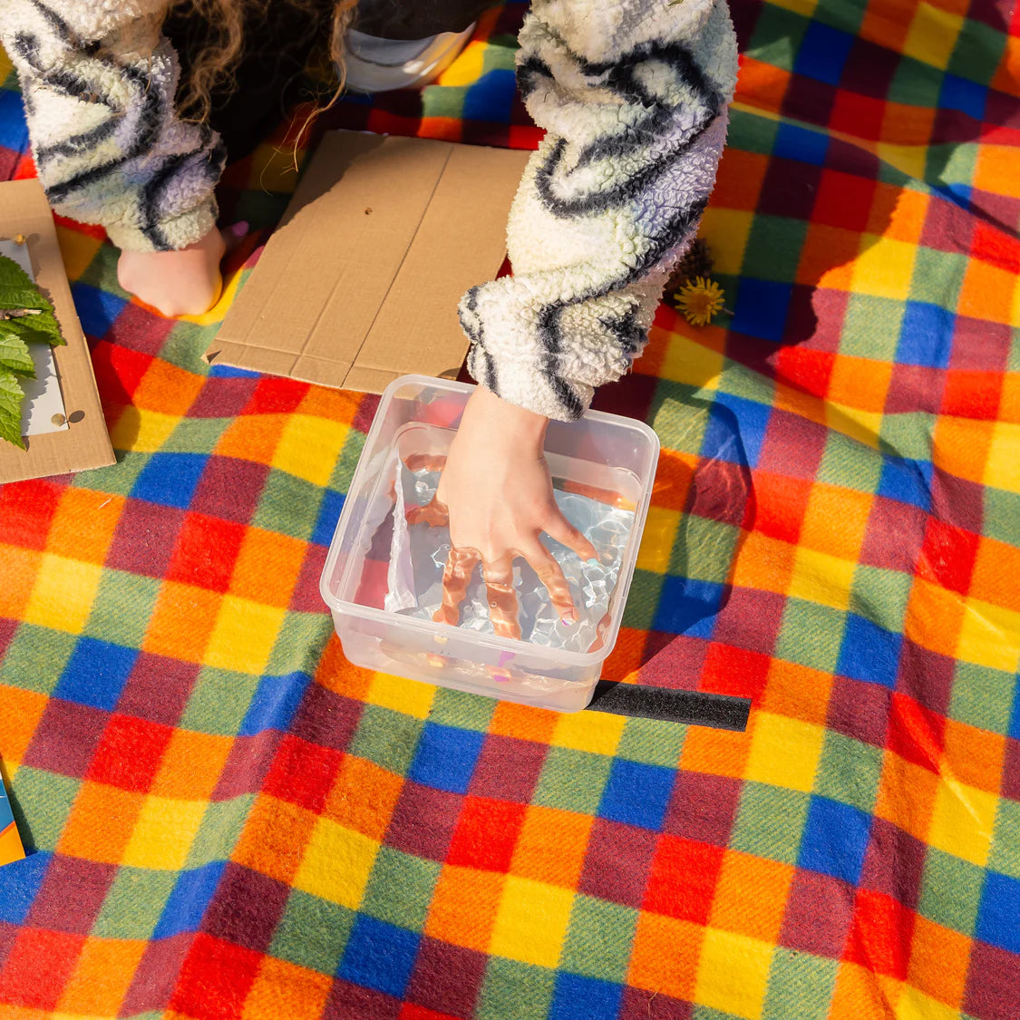 Child's hand in a clear container of water on a colorful checkered blanket