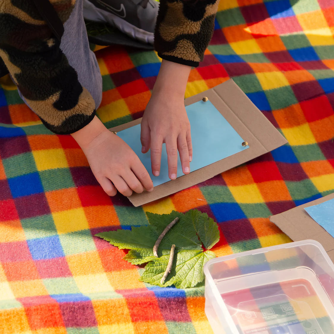 Child's hands interacting with a leaf and caterpillar on a colorful checkered blanket