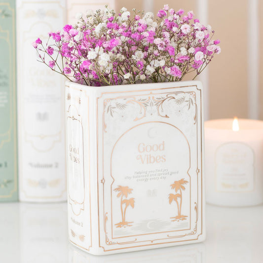 Decorative book shaped vase with 'Good Vibes' text and pink flowers on a light background